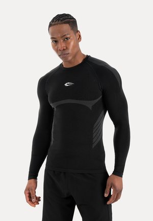 Atletisk mand iført sort langærmet kompressionsshirt med subtile teksturmønstre og matchende sorte bukser, stående mod en ensfarvet baggrund.