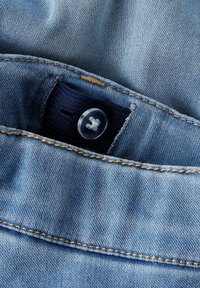 Nærbilde av midjebånd på blå denimjeans med justerbart elastisk bånd og gjennomsiktig plastknapp for størrelsesjustering.