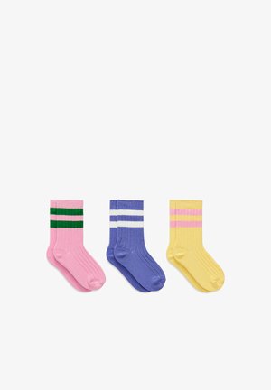 Trois paires de chaussettes côtelées rose avec des rayures vertes, bleu avec des rayures blanches et jaune avec des rayures roses, disposées côte à côte sur fond blanc.