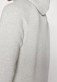 Hoodie gris clair avec un tissu doux et texturé. Il présente une poche kangourou et une capuche à cordon, montrant un gros plan de la couture latérale.