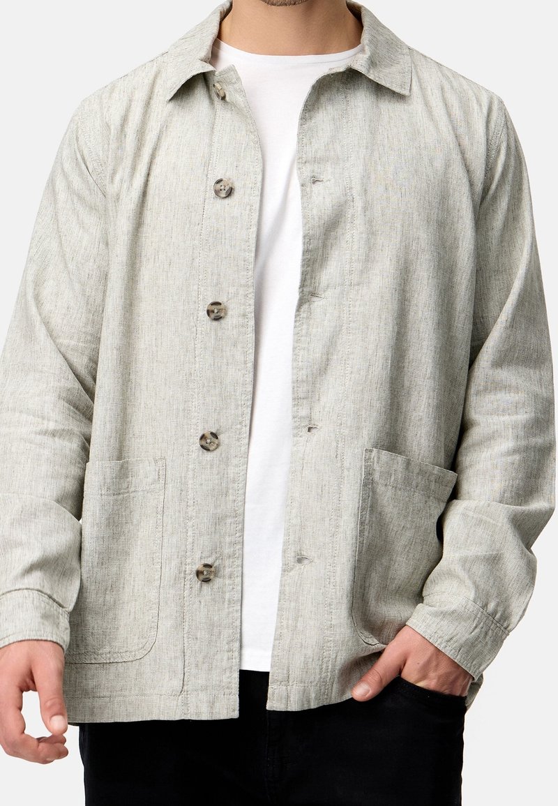 Homme portant une veste boutonnée gris clair sur une chemise blanche, une main dans la poche d'un pantalon noir, visible du cou aux hanches.