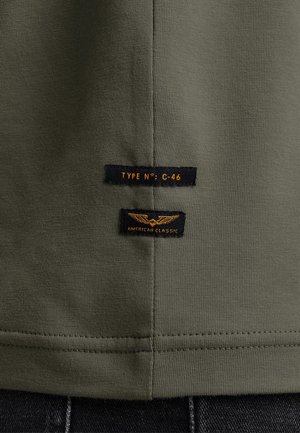 Olivgrünes T-Shirt mit zwei schwarzen Stofflabels mit orangefarbigem Text. Eines der Labels trägt die Aufschrift "TYPE N°: C-46", das andere "AMERICAN CLASSIC."
