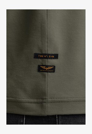 Olivgrünes T-Shirt mit zwei schwarzen Stofflabels mit orangefarbigem Text. Eines der Labels trägt die Aufschrift "TYPE N°: C-46", das andere "AMERICAN CLASSIC."