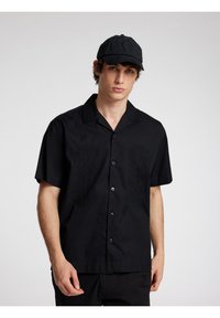 Camicetta nera a maniche corte con colletto, caratterizzata da ricami texture sul davanti. Abbinata a un cappellino nero coordinato.