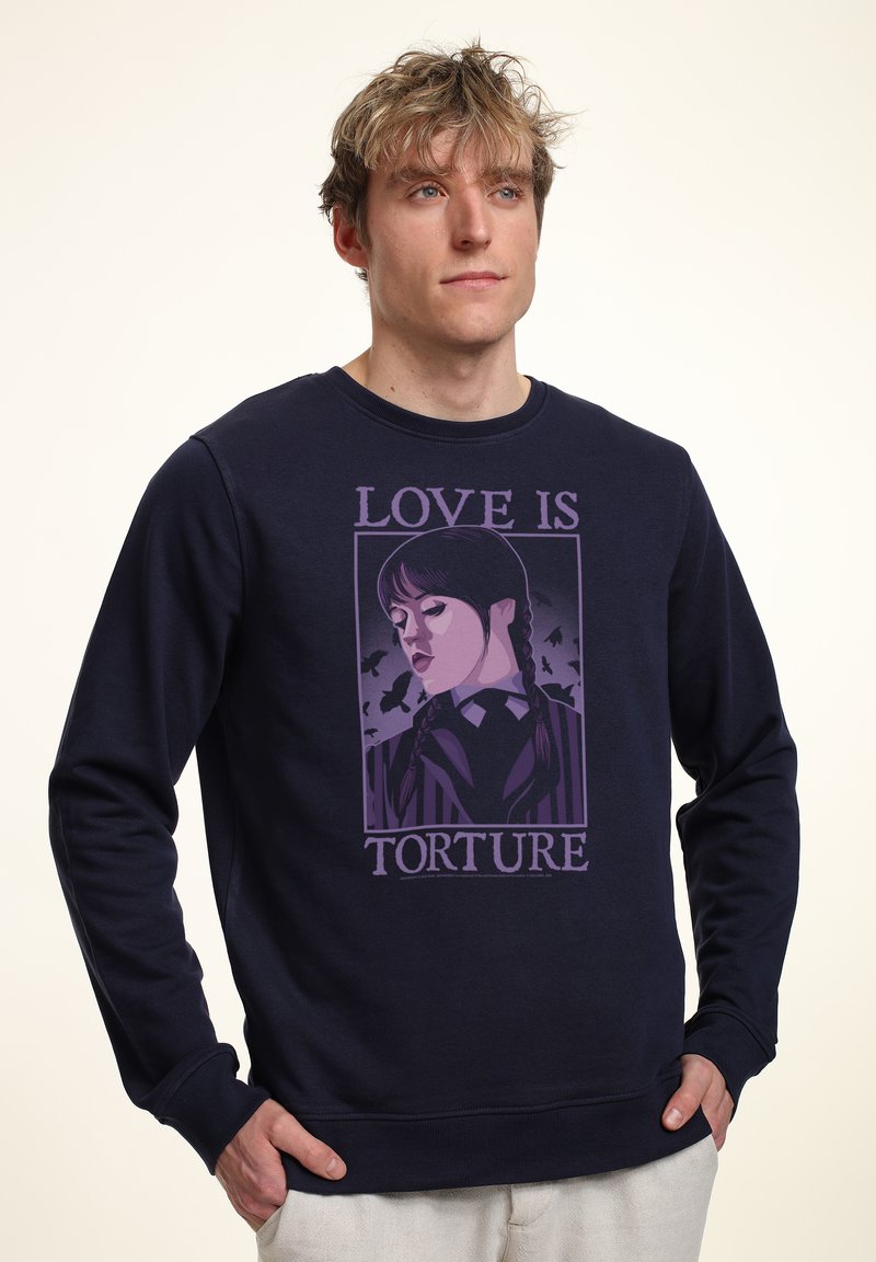 Uomo biondo che indossa una felpa blu navy con una grafica viola di una donna con capelli intrecciati e la scritta "L'AMORE È TORTURA" sul davanti.