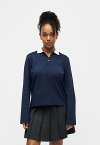 Jeune femme aux cheveux bouclés attachés en queue de cheval haute, portant un polo à manches longues bleu marine avec un col blanc et une jupe plissée sombre.