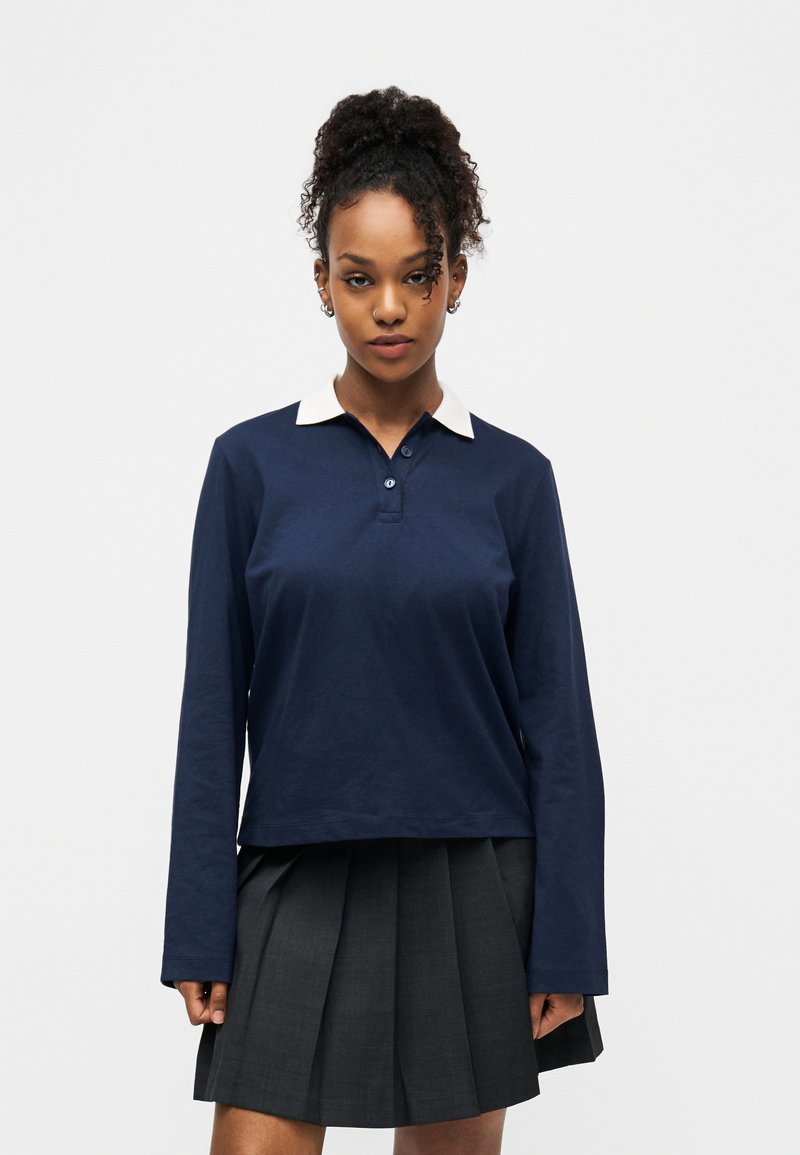 Jeune femme aux cheveux bouclés attachés en queue de cheval haute, portant un polo à manches longues bleu marine avec un col blanc et une jupe plissée sombre.