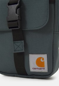Saco de tecido verde-escuro com uma alça de fivela preta. Apresenta um rótulo cinza com um logótipo laranja e texto vermelho, mostrando um design simples e compacto.