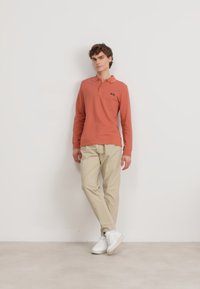 Polo a maniche lunghe corallo con colletto bianco, abbinato a pantaloni beige e sneaker bianche, esposto su uno sfondo neutro.