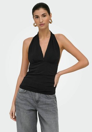 Kobieta z ciemnymi włosami w czarnym topie z odkrytymi plecami i szarych jeansach, pozująca z jedną ręką na biodrze na jasnym, jednolitym tle.