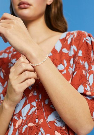 Weißes Perlenarmband mit kleinen, runden Perlen an einer zarten Kette, am Handgelenk getragen. Der Hintergrund zeigt ein rotes, blumengemustertes Oberteil.