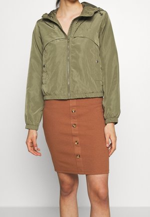 Kobieta w oliwkowo-zielonej bluzie z kapturem i zamkiem, oraz brązowej spódnicy z guzikami z przodu, stojąca na jednolitym jasnym tle.