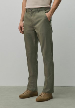 Olívazöld egyenes szárú nadrágot és bézs színű velúr sportcipőt viselő férfi, kezét a zsebében tartva egy sima háttér előtt áll.