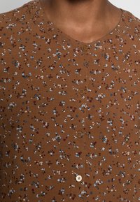 Camicia a maniche corte marrone con bottoni, caratterizzata da piccoli fiori blu e rossi su un tessuto testurizzato, con cinque bottoni lungo il collo davanti.