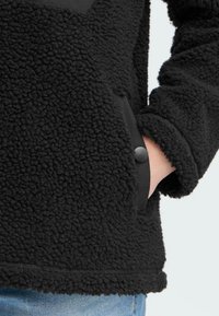 Zwarte fleecetopper van textuurachtig, donzig materiaal. Heeft een voorzak met een zwarte knopaccent en een glad, contrasterend paneel.