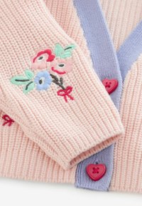 Rozā adīts kardigans ar pasteļziliem maliņiem, izšūti krāsaini ziedi uz piedurknes un sarkanas sirds formas pogas.