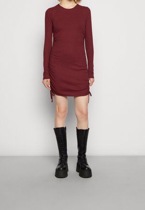Burgunderfarbenes langärmliges Kleid mit seitlichen Raffungselementen, kombiniert mit knielangen schwarzen Schnürstiefeln. Glattes Material, figurbetonte Silhouette.