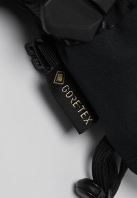 Gore-Tex-label bevestigd aan een zwarte stof, met een gestructureerd oppervlak, metallic accenten en een plastic clipbevestiging.