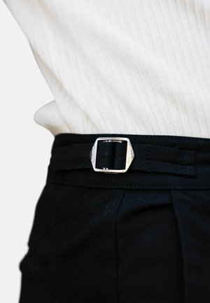 Cinturón de tela negra con hebilla cuadrada de plata, llevado a la altura de la cintura sobre una camiseta de canalé clara. La hebilla lleva grabado el nombre de la marca.