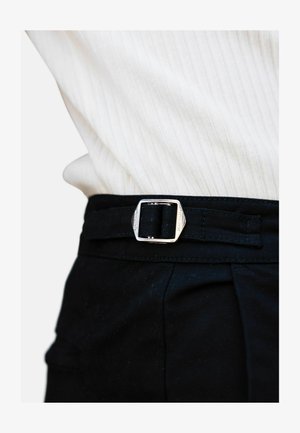 Black fabric belt with a silver square buckle, worn at waist level over a light ribbed top. Buckle features engraved brand name.