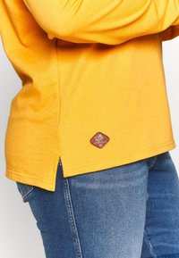Shirt gialla a maniche lunghe con scollatura rotonda, fessure laterali e un'etichetta con logo marrone nella parte inferiore. Tessuto morbido realizzato in tessuto leggero.