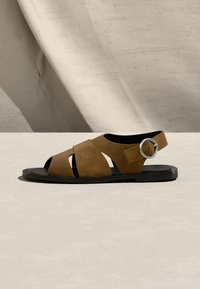 Καφέ σουέντ σανδάλι με ανοιχτό δάχτυλο, φαρδιές λουρίδες και ασημένια στρογγυλή αγκράφα σε μαύρη επίπεδη σόλα σε μπεζ φόντο.