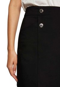 Jupe noire avec une texture lisse, taille haute, dotée de deux boutons métalliques à l'avant et d'une main reposant sur le tissu sur le côté.