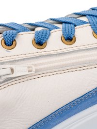 White sneaker side with blue laces threaded through brass eyelets and a white zipper running parallel below the lace row.