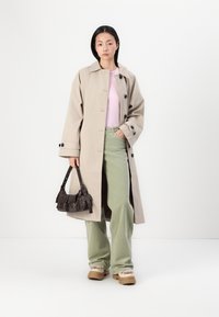 Manteau long beige avec des boutons noirs, haut rose pâle, pantalon vert évasé et sac à main marron. Les chaussures présentent une base tan avec des accents en fourrure.