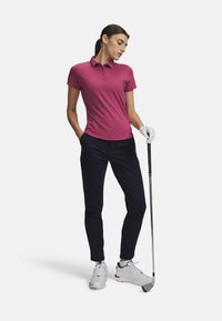 Ροζ πόλο μπλούζα με κοντό μανίκι και γιακά, συνδυασμένη με ναυτικό μπλε παντελόνι, λευκά παπούτσια του γκολφ και ένα γκολφ κλαμπ στο έδαφος. Γάντι του γκολφ σε ένα χέρι.