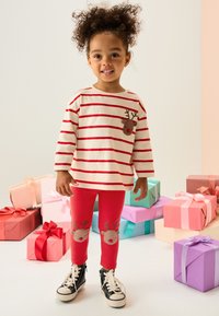 Maglia a maniche lunghe a righe rosse e bianche con toppa a forma di renna; leggings rossi con facce di renna sulle ginocchia; scarpe da ginnastica nere.