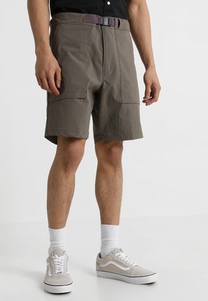 Mand iført taupe shorts med forlommer, et lilla bælte, hvide crewstrømper og lysegrå sneakers mod en hvid baggrund.