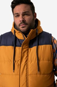 Pufferväst i senapsgul med marinblå detaljer, med hög krage, dragsko och quiltade sektioner. Mjuk, isolerad tyg.