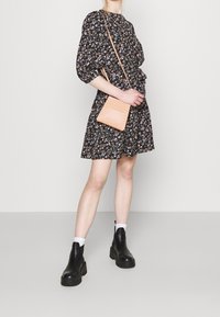 Vrouw in zwarte jurk met bloemenpatroon en zwarte enkellaarsjes die een tan schoudertas vasthoudt tegen een effen achtergrond.