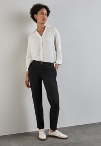 Vit, lätt skjorta med knappar; svarta slim-fit byxor; vita sneakers med beige detaljer; stående pose mot en neutral bakgrund.