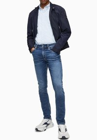 Námořnická lehká bunda, světle modrá košile s knoflíky, modré džínové kalhoty a šedé tenisky. Bunda má zipové kapsy a úzký design.