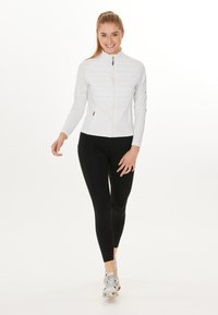 Vit zip-jacka med vadderad front, långa ärmar och en figursydd design, tillsammans med svarta leggings och löparskor.