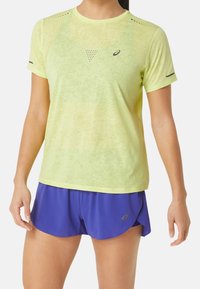 Jasnożółta, krótka sportowa koszulka z fakturowanym wykończeniem i perforowanymi detalami, w połączeniu z fioletowymi siateczkowymi szortami z rozcięciami po bokach.