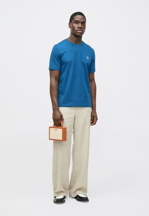 Modré bavlněné tričko s malým vyšitým vzorem, doplněné béžovými širokými kalhotami a černými botami s bílými detaily, držící malou hnědou kabelku.