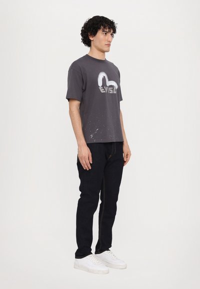 Camiseta gris de manga corta con un logo blanco y detalles de salpicaduras, combinada con jeans oscuros y zapatillas blancas.