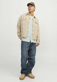 Veste en denim beige sur une chemise à carreaux bleu clair, jeans bleus et bottes beiges, avec une casquette grise arborant un logo. Design simple et coupe décontractée.