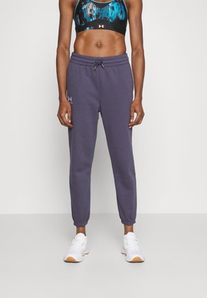 Woman wearing navy blue Under Armour joggers, white sneakers, and a blue patterned sports bra, standing against a plain white background.