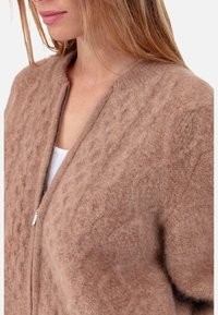 Brauner, gestrickter Cardigan aus weichem Material. Verfügt über ein strukturiertes Muster, einen Reißverschluss und einen gerippten Ausschnitt.