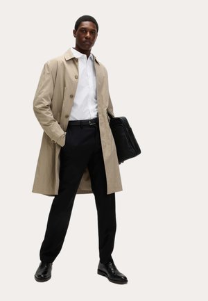 Mężczyzna ubrany w beżowy płaszcz przeciwdeszczowy, białą koszulę, czarne spodnie i czarne buty, trzymający czarną teczkę, stojący na tle jednokolorowego tła.