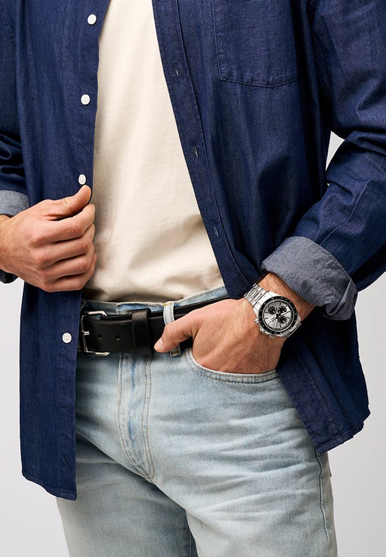 Granatowa koszula z guzikami, biała koszulka pod spodem, jasnoniebieskie dżinsy, czarny pasek oraz srebrny zegarek z czarną tarczą.