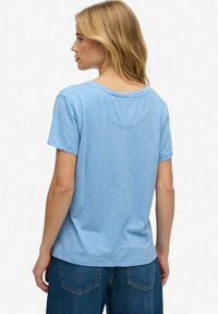 Vrouw met blond haar die een lichtblauwe T-shirt met korte mouwen en donkerblauwe jeans draagt, gezien van achteren tegen een witte achtergrond.