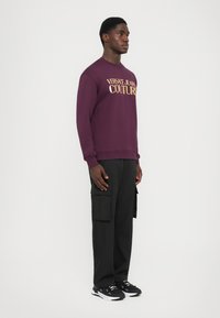 Felpa viola con "VERSACE JEANS COUTURE" in lettere dorate. Pantaloni cargo neri con tasche. Scarpe sportive nere. Silhouette slim fit.