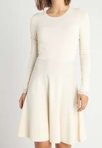 Långärmad klänning i krämfärg med åtsittande bodice och utsvängd kjol. Har texturerad tyg och spetsdetaljer vid ärmsluten.