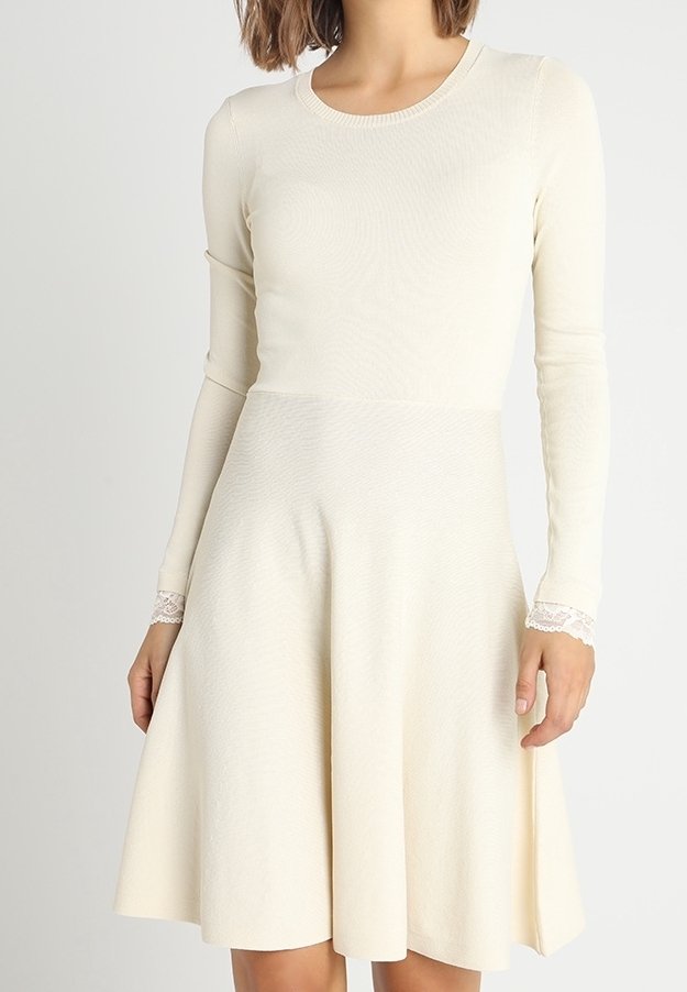 Långärmad klänning i krämfärg med åtsittande bodice och utsvängd kjol. Har texturerad tyg och spetsdetaljer vid ärmsluten.