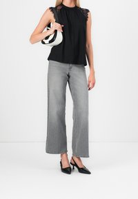 Donna che indossa un top senza maniche nero con pizzo, jeans grigi a gamba larga, tacchi neri con cinturino alla caviglia, e tiene in mano una borsa a spalla bianca e nera.
